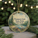 A round ornament with a scenic lake and forest landscape, featuring pine trees and a cabin, is displayed in front of a blurred background with Christmas lights.
