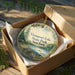 A decorative ornament in a wooden box, featuring a scenic landscape with pine trees, a lake, and a cabin, with the text "Thomson's Lake State Park, New York" printed on it.