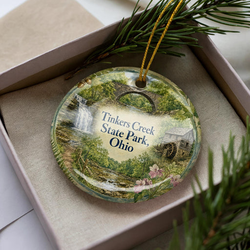 A decorative ornament with a scenic image of Tinkers Creek State Park, Ohio, displayed in a box with pine branches.