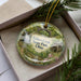 A decorative ornament with a scenic image of Tinkers Creek State Park, Ohio, displayed in a box with pine branches.