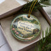 A decorative ornament in a box, featuring a scenic landscape with a waterfall, pine trees, and a cabin, along with the text "Tomlinson Run State Park, West Virginia".