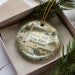 A decorative ornament in a box, featuring a scenic landscape with a tower, trees, and a body of water.