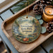 A decorative ornament with a scenic landscape of Townshend State Park, Vermont, featuring a waterfall, pine trees, and a bridge. The ornament is displayed on a wooden tray, accompanied by pine cones and a lit candle.