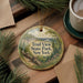 A round ornament with a scenic landscape of Trail State Park, New York, featuring a bridge and pine trees, is displayed on a wooden surface alongside a cup of coffee and some papers.