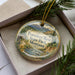 A decorative ornament in a box, featuring a scenic landscape with mountains, water, and pine trees.