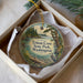 A circular ornament with a scenic landscape of a lake, trees, and a bird, displayed in a box with a white tissue paper lining.