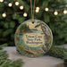 A round ornament with a scenic image of a park, featuring a bird, trees, and a body of water, hanging in front of a Christmas tree with lights.