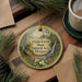 A circular ornament with a nature scene and text, placed on a wooden surface next to a cup of coffee and pine branches.