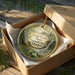 A decorative ornament in a wooden box, featuring a natural scene with ferns, flowers, and a waterfall.