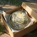 A decorative ornament in a box, featuring a scenic landscape with a waterfall and pine trees, with the text "Tumalo State Park, Oregon" printed on it.