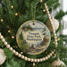 A round ornament hanging on a Christmas tree, featuring a scenic landscape with a lake, mountains, and pine trees.