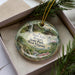 A decorative ornament in a box, featuring a scenic image of Twin Falls Resort State Park in West Virginia.