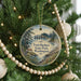 A round ornament hanging on a Christmas tree, featuring a scenic landscape with a lighthouse and a river.