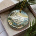 A decorative ornament in a box, featuring a lighthouse and a scenic landscape.