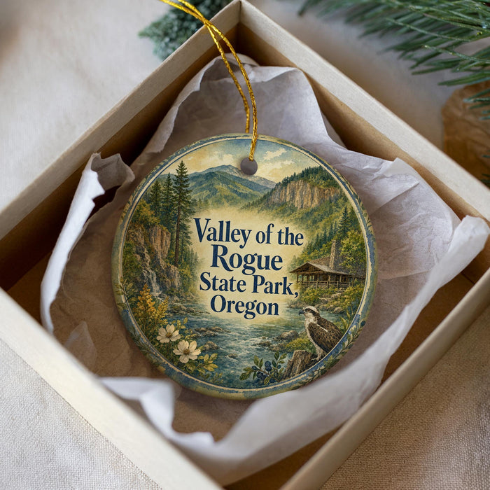 A decorative ornament in a box, featuring a scenic landscape with mountains, trees, and a bird, along with the text "Valley of the Rogue State Park, Oregon".