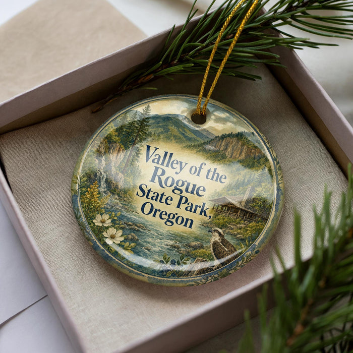 A decorative ornament with a scenic landscape of mountains, trees, and a bird, displayed in a box with pine branches.