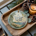 A decorative ornament with a scenic beach landscape, featuring a lifeguard tower, is displayed on a wooden tray alongside pine cones and a lit candle.