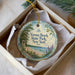 A round ornament with a scenic beach landscape and text, placed in a box with a white tissue paper lining.