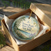 A decorative ornament in a cardboard box, featuring a scenic image of Verona Beach State Park in New York.