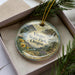 A decorative ornament with a landscape scene featuring mountains, pine trees, and a waterfall, placed inside a box with pine branches.