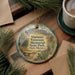 A decorative ornament with a picture of a memorial park, specifically Vietnam Veterans Memorial in New Mexico, is displayed on a wooden surface alongside a cup of coffee and some pine branches.