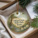 A decorative ornament with a nature scene, featuring a bird, a deer, and a stream, is displayed in a box with pine branches.