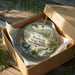 A decorative ornament in a box, featuring a scenic landscape with a waterfall and pine trees, with the text "Wadleigh State Park, New Hampshire" printed on it.