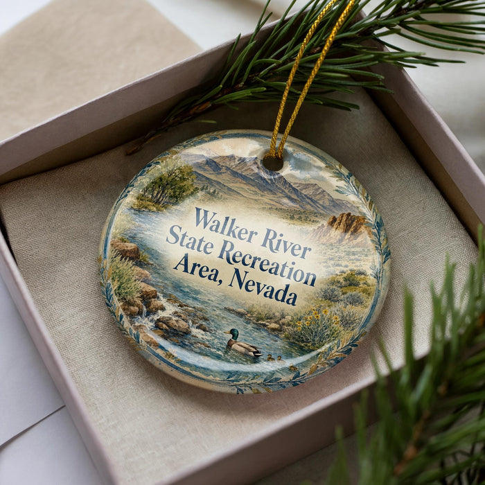 A decorative ornament with a landscape scene featuring a river, mountains, and wildlife, including a duck, is displayed in a box with pine branches.