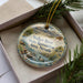 A decorative ornament with a landscape scene featuring a river, mountains, and wildlife, including a duck, is displayed in a box with pine branches.