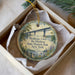 A decorative ornament in a box, featuring a scenic image of a bridge over a river surrounded by trees and flowers.