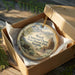 A decorative ornament in a wooden box, featuring a scenic landscape with mountains and a chimney, is placed on a wooden surface.