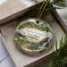 A decorative ornament in a box, featuring a scenic landscape with a waterfall and pine trees.