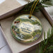 A decorative ornament in a box, featuring a scenic landscape with a bridge, trees, and a river.