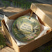 A decorative ornament in a box, featuring a scenic landscape with a waterfall and pine trees, with the text "Watoga State Park, West Virginia" prominently displayed.