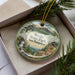 A decorative ornament with a scenic landscape of Weeks State Park in New Hampshire, featuring a mountain, waterfall, and pine trees.