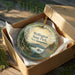 A round ornament in a box, featuring a scenic landscape with a lake, mountains, and pine trees.