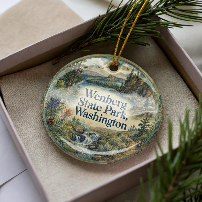 A decorative ornament in a box, featuring a scenic landscape with mountains, waterfalls, and pine trees.