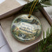 A decorative ornament in a box, featuring a scenic landscape with mountains, waterfalls, and pine trees.