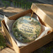 A decorative ornament in a wooden box, featuring a scenic landscape with a river, trees, and a bridge.