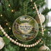 A round ornament hanging on a Christmas tree, featuring a scenic landscape with a river, bridge, and trees.
