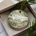 A decorative ornament in a box, featuring a scenic landscape with a beach, a house, and a body of water.