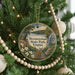 A round ornament hanging on a Christmas tree, featuring a scenic landscape with a waterfall and a deer.