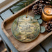 A wooden tray with a decorative ornament featuring a turtle and a pine tree, surrounded by pine cones and a lit candle.