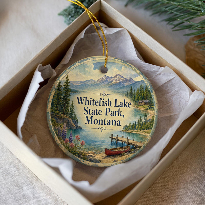 A decorative ornament in a white box, featuring a scenic landscape with mountains, pine trees, and a lake with a boat on the shore.