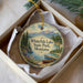 A decorative ornament in a white box, featuring a scenic landscape with mountains, pine trees, and a lake with a boat on the shore.