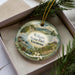 A decorative ornament with a scenic landscape of Wilgus State Park, Vermont, displayed in a box with pine branches.