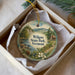 A decorative ornament in a white box, featuring a scenic landscape with mountains, a river, and a cabin.