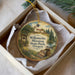 A round ornament with a painting of a scenic landscape, including a bell tower, trees, and a river, is displayed in a white box.