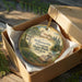 A wooden ornament in a box, featuring a scenic landscape with a house, trees, and a river.