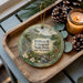 A decorative ornament featuring a scenic landscape of William B. Umstead State Park in North Carolina, with a deer and a stream, is displayed on a wooden tray alongside pine cones and a lit candle.